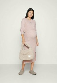 Robe en tricot à manches longues, couleur violette, avec des détails de boutons et une fente sur le côté. Le modèle tient un sac à main moelleux de couleur gris clair et porte des mocassins beiges.