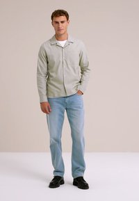 Svetlozelena srajca z dolgimi rokavi, ena žep na prsih, v kombinaciji s svetlo modrimi kavbojkami in črnimi čevlji, stoji ob nevtralni ozadju.