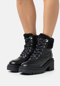 Bottes de cheville en cuir noir avec un col en fourrure noire texturée, un design robuste à lacets, des éléments en métal argenté et une semelle plateforme épaisse et robuste.