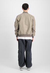 Khaki bomber-jakk sileda tekstuuriga; soonikuga krae; passitud varrukad; kantud valge särke ja ühendatud lahtiste hallide pükstega.