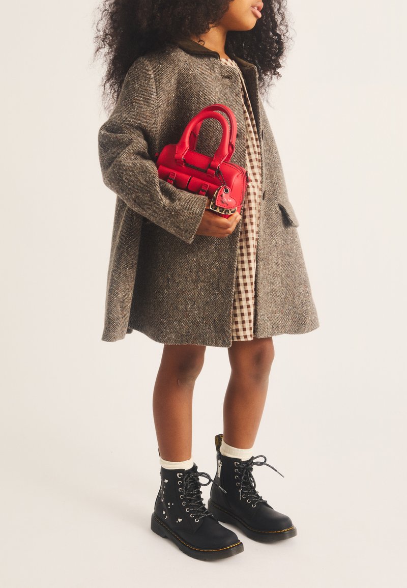 Jeune fille aux cheveux bouclés portant un manteau gris, une robe à carreaux, des bottes noires à lacets, tenant un petit sac à main rouge.