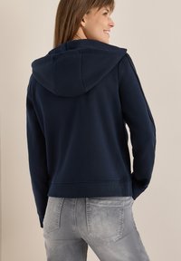 Donna che sorride di lato, indossando una felpa con cappuccio blu navy e jeans grigi chiari, in piedi contro un muro beige.