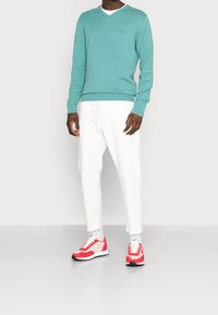 Turkusowy sweter w serek, białe spodnie z postrzępionymi mankietami oraz czerwone buty sportowe z białymi i czarnymi akcentami.