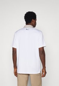 Camisa polo branca com colarinho bege, mangas curtas e um logo preto da Nike nas costas. Apresenta fendas laterais para um movimento aprimorado.