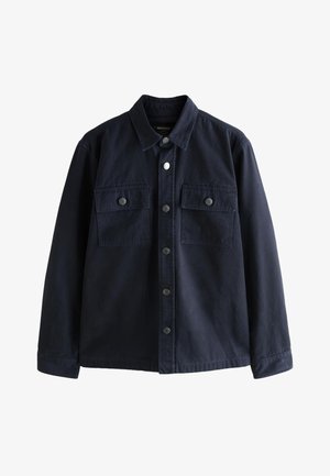 Junger Mann, der auf einer Kopfsteinpflasterstraße steht, trägt eine dunkelblaue Jacke, eine graue Kapuzenjacke, beige Hose und blaue Turnschuhe und schaut nach links.