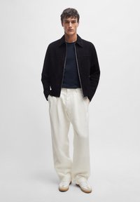 Sort ruskind bomberjakke over en marineblå top, parret med løse hvide bukser og hvide sneakers med tan accenter. Enkel, moderne design.