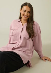 Camisa de botones de gran tamaño en rosa claro con dos bolsillos frontales, mangas largas y un dobladillo curvado, combinada con una falda negra. Textura de tela suave.