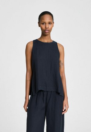 Sötét bőrű, rövid hajú nő ujjatlan fekete felsőt és hozzá illő, bő szárú nadrágot visel, egyszerű világos háttér előtt áll.