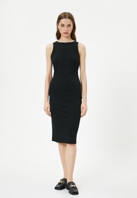 Schwarzes ärmelloses Midikleid aus glattem Stoff. Figurbetonte Silhouette mit Rundhalsausschnitt, kombiniert mit schwarzen strukturierten Slippern mit goldenen Akzenten.