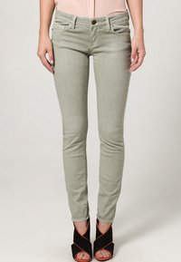 Jeans ajustados de color verde claro con una textura suave, cinco bolsillos, un botón de metal y detalles de costura sutiles, combinados con sandalias de tacón negra.