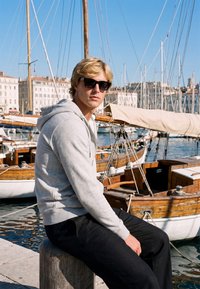 Jeune homme portant des lunettes de soleil et un sweat à capuche gris assis sur un quai à côté de voiliers en bois dans une marina ensoleillée avec des bâtiments en arrière-plan.