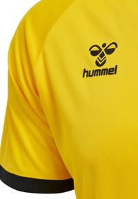 Gul sportstrøje lavet af glat, syntetisk stof. Har sorte accenter på ærmerne og "hummel" logoet trykt på den øverste venstre side.