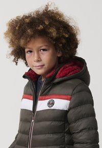 Niño con pelo rizado que lleva una chaqueta acolchada verde, roja y blanca con capucha forrada de piel roja, de pie contra un fondo liso.