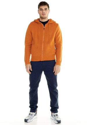 Un homme se tient debout, de face, sur un fond blanc. Il porte un sweat à capuche orange zippé, un pantalon bleu marine et des baskets blanches.