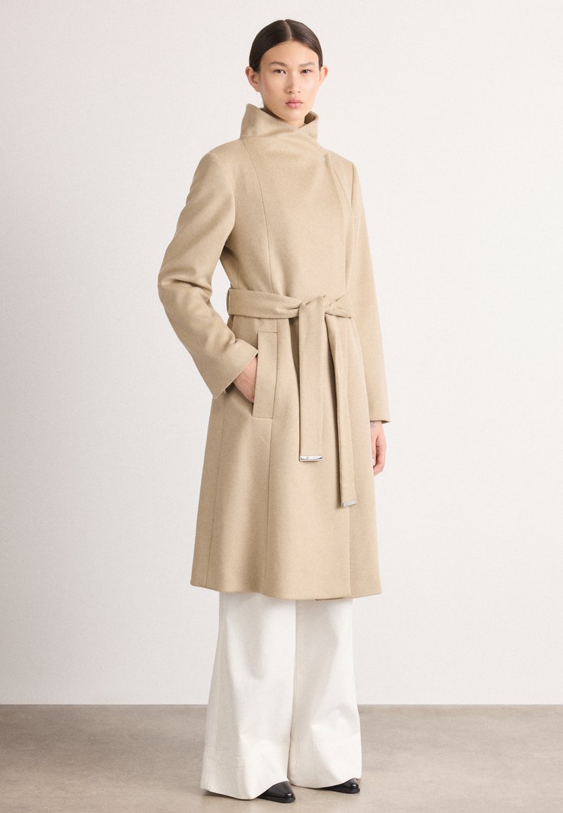 Abrigo de lana beige con cuello alto, cintura ceñida y bolsillos laterales. Lleva sobre unos pantalones blancos anchos, con una textura suave y un diseño minimalista.