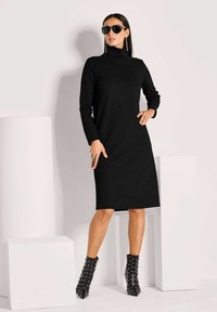 Frau, die ein schwarzes langärmeliges Rollkragenkleid, mit Nieten verzierte schwarze Knöchelschuhen, Sonnenbrille und langen Ohrringen trägt, posiert mit der Hand auf der Hüfte.