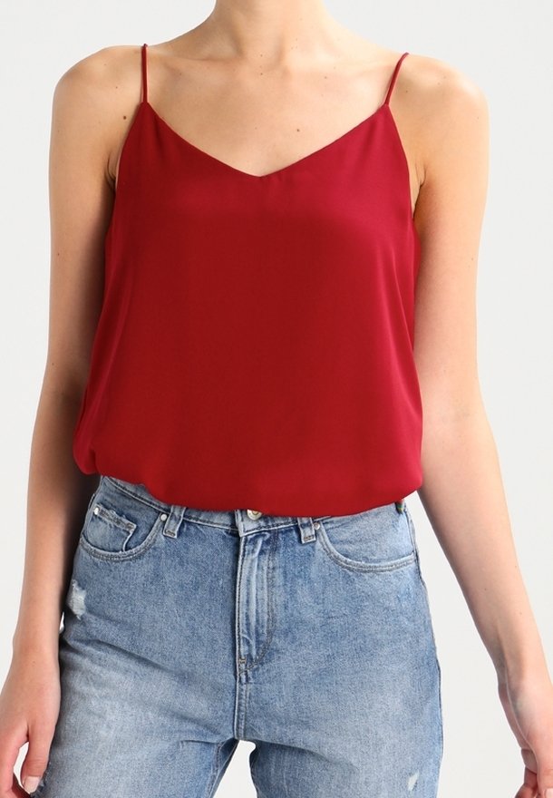 Femme portant un haut sans manches rouge avec de fines bretelles et un jean bleu clair taille haute légèrement usé.