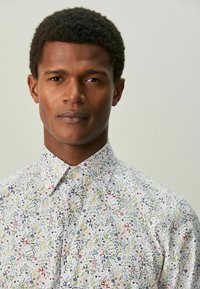 Valge nööpidega särk, mis on kaunistatud värvikireva lillemustriga, millel on rohelised, punased, sinised ja kollased aktsendid. Sile kangapind.