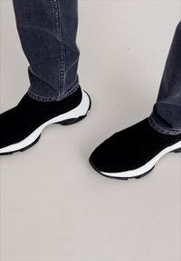 Fekete hálós slip-on sportcipő fehér talppal, letisztult dizájnnal és texturált anyaggal. Feszes szabású farmernadrággal párosítva.