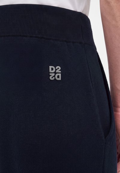 Gros plan d'une personne portant un pantalon bleu marine foncé avec une ceinture côtelée et un logo métallique D2 sur la partie supérieure de la cuisse près d'une poche.