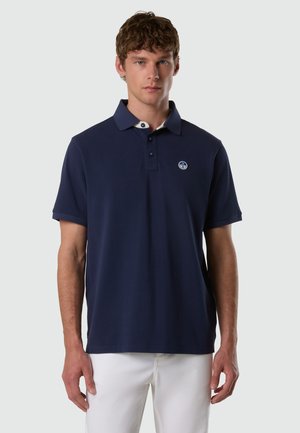Modello maschile che indossa una polo blu navy a maniche corte con colletto interno bianco e piccolo logo rotondo, abbinata a pantaloni bianchi, rivolto verso l'obiettivo.
