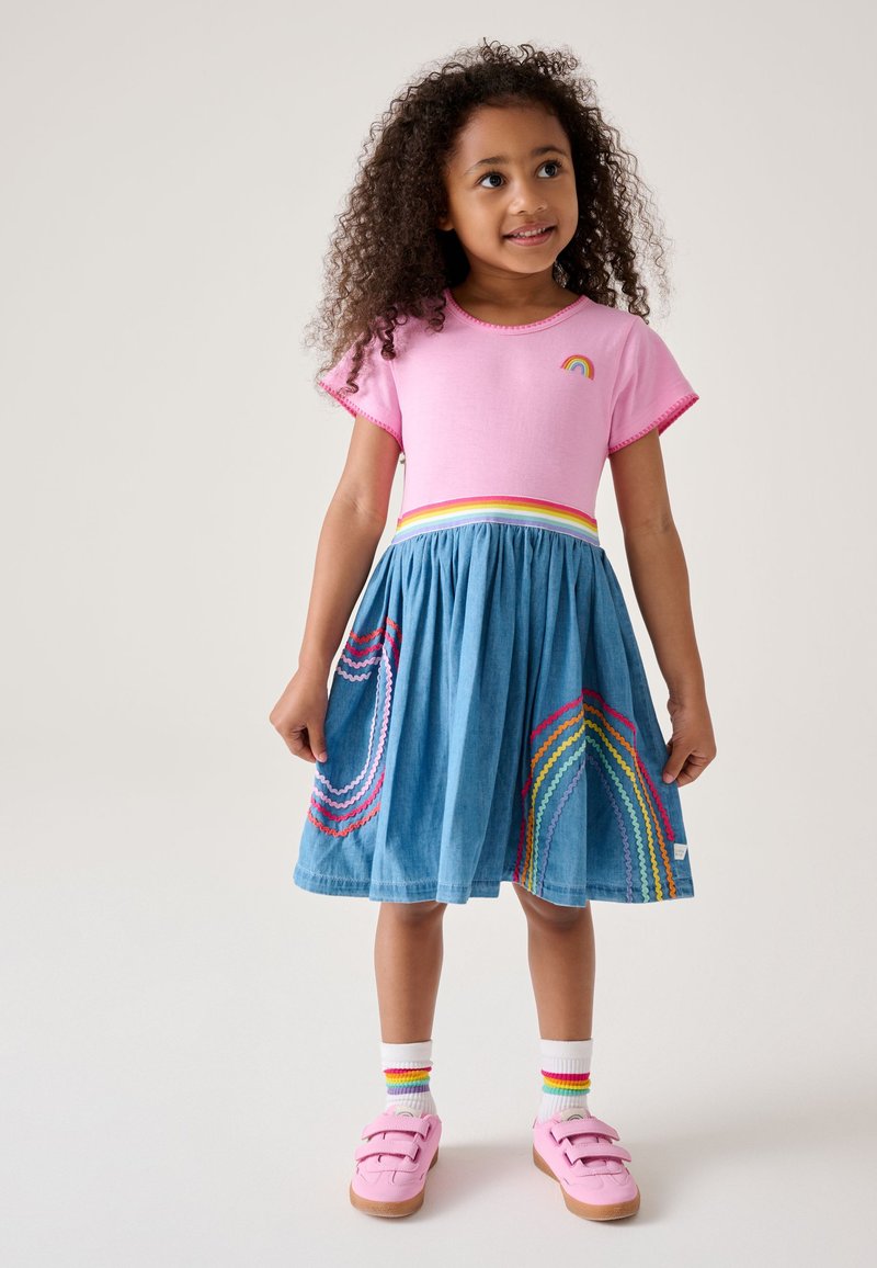 Ragazza giovane con capelli ricci, indossa una maglietta rosa con dettaglio arcobaleno, gonna blu con motivi arcobaleno, calzini bianchi con arcobaleno e scarpe rosa.