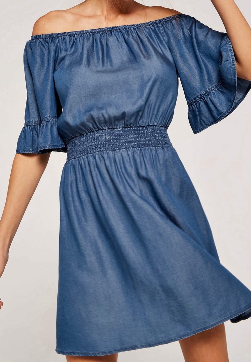 Femme portant une robe bleue décolletée avec une taille élastique et des manches courtes évasées, montrée des épaules jusqu'au milieu de la cuisse.