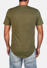 T-shirt verde oliva a maniche corte con orlo arrotondato e texture liscia, abbinato a jeans in denim chiaro. Design semplice, senza grafiche visibili.
