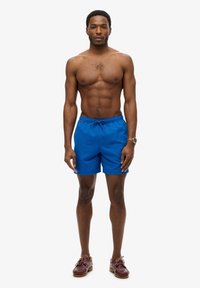 Pantaloni da bagno blu con vita elastica regolabile, tasche laterali e vestibilità ampia, abbinati a scarpe marroni senza lacci.