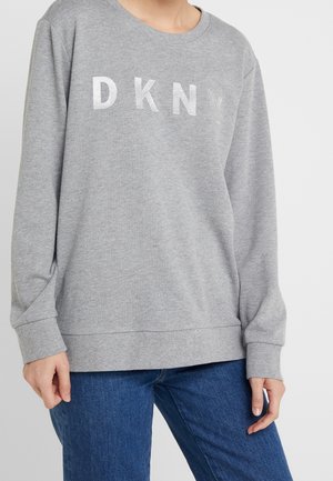 Persoon die een grijze DKNY-trui en blauwe spijkerbroek draagt, gezien van schouders tot heupen tegen een effen achtergrond.