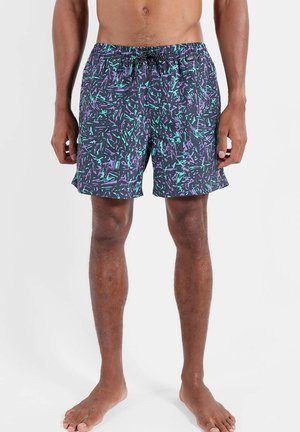 Homme portant un short de bain aux motifs abstraits multicolores noir, violet et sarcelle, debout pieds nus devant un fond blanc uni.