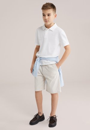 Jeune garçon debout, les mains dans les poches, portant un polo blanc, un short beige, des chaussures noires, et une chemise rayée bleue nouée autour de la taille.