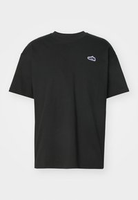 Camiseta de algodón negra con cuello redondo y mangas cortas. Presenta un pequeño logo de zapatilla azul y blanco en el área del pecho izquierdo.