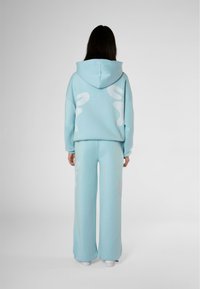 Felpa con cappuccio azzurro chiaro e pantaloni della tuta a gamba larga coordinati, caratterizzati da grandi disegni astratti bianchi sul retro. Texture morbida, vestibilità casual.