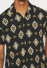 Kortärmad skjorta i svart tyg med ett geometriskt mönster i gult, vitt och krämiga nyanser, knäppning fram och kampkrage.
