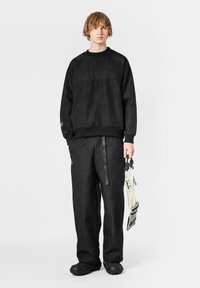 Sudadera negra con patrón texturizado y logo, combinada con pantalones negros texturizados a juego. El modelo sostiene un accesorio blanco con forma de correa.