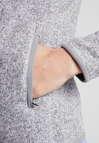 Grå stickad sweatshirt med ett texturerat mönster och grå kant, med en ficka med dragkedjestängning på sidan.