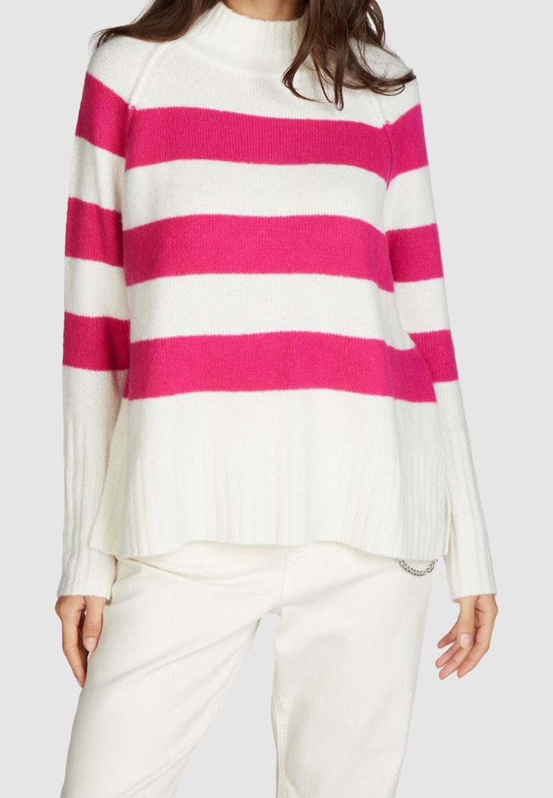 Pull en maille blanche avec de larges rayures rose fuchsia, doté d'un col montant, d'un ourlet côtelé et de fentes latérales, associé à un pantalon de couleur claire.