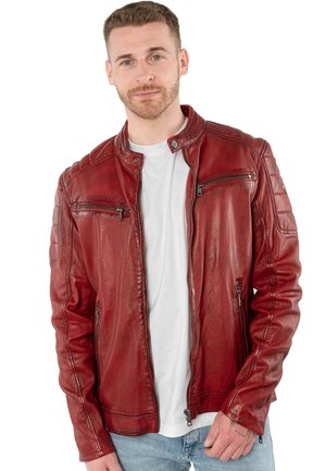 Mann mit kurzen Haaren und Bart, der eine rote Lederjacke über einem weißen T-Shirt und blauen Jeans trägt, steht vor einem weißen Hintergrund.