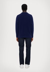 Navy knit sweater with a high collar, ribbed hem, and raglan sleeves, paired with dark jeans and white sneakers. Back view of the outfit.