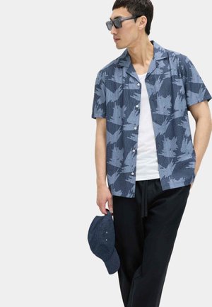 Mann trägt blau gemustertes Kurzarmhemd über weißem T-Shirt und schwarze Hose, hält dunkelblaue Kappe in der Hand und trägt schwarze Sonnenbrille.