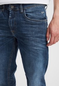 Denimjeans i mørk blå med en vanlig passform, som har fem lommer, synlige sømmer og en liten logo på baklommen.