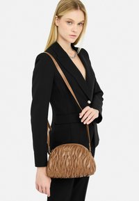 Sac en cuir marron avec une texture matelassée et une forme arrondie, doté d'une seule bandoulière et d'un détail en logo métallique.