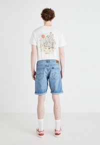 Ανοιχτόχρωμο γραφικό t-shirt με σχέδιο κάκτου στο πίσω μέρος, σε συνδυασμό με μπλε τζιν σορτς και κόκκινα αθλητικά παπούτσια Nike.