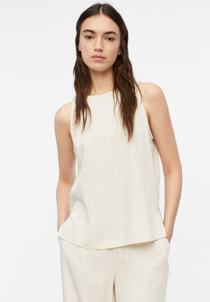 Kobieta z długimi brązowymi włosami, ubrana w białą bluzkę bez rękawów i białe spodnie, stoi z rękami w kieszeniach na jednolitym tle.