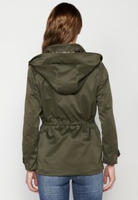 Kobieta z długimi brązowymi włosami, ubrana w oliwkową zieloną kurtkę z kapturem i niebieskie dżinsy, widziana z tyłu na tle białego tła.