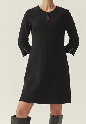 Femme portant une robe noire mi-longue avec manches trois-quarts, fentes sur le devant et petit fermoir au niveau du col.
