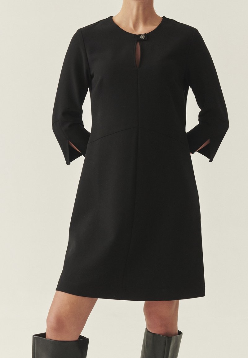 Femme portant une robe noire mi-longue avec manches trois-quarts, fentes sur le devant et petit fermoir au niveau du col.