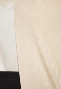 Cardigan a coste di un beige chiaro realizzato in materiale morbido, con una finitura testurizzata. Indossato sopra una maglietta color crema e un indumento scuro sottostante.