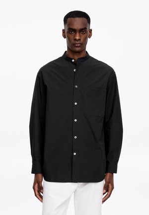 Mand iført en sort langærmet skjorte med hvide knapper og en brystlomme, kombineret med hvide bukser foran en ensfarvet hvid baggrund.